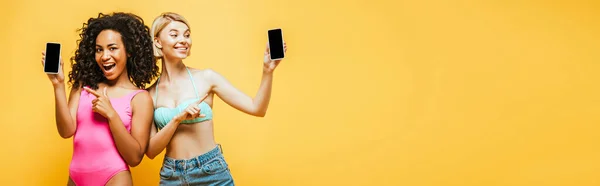 Сайт заголовок молодых межрасовых женщин в летнем наряде указывая на смартфоны с чистым экраном изолированы на желтый — стоковое фото