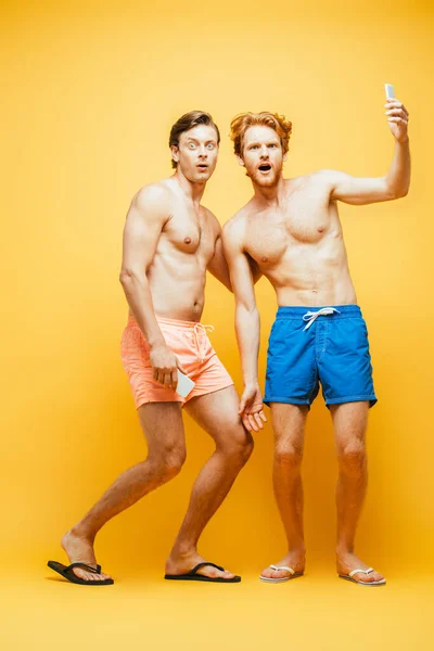 Vista completa de dos hombres sin camisa en pantalones cortos haciendo muecas mientras mira a la cámara y toma selfie en el teléfono inteligente en amarillo - foto de stock