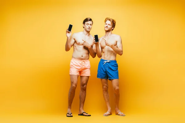 Vista a tutta lunghezza di due uomini senza maglietta in pantaloncini puntati con le dita sugli smartphone su giallo — Foto stock