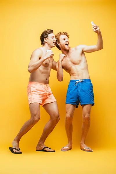 Vista completa de dos hombres emocionados haciendo muecas mientras muestran los puños apretados y tomando selfie en el teléfono inteligente en amarillo - foto de stock