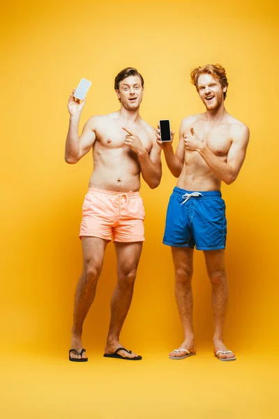 Visão de comprimento total de dois amigos sem camisa em shorts mostrando o polegar para cima e apontando com o dedo enquanto segura smartphones no amarelo — Fotografia de Stock