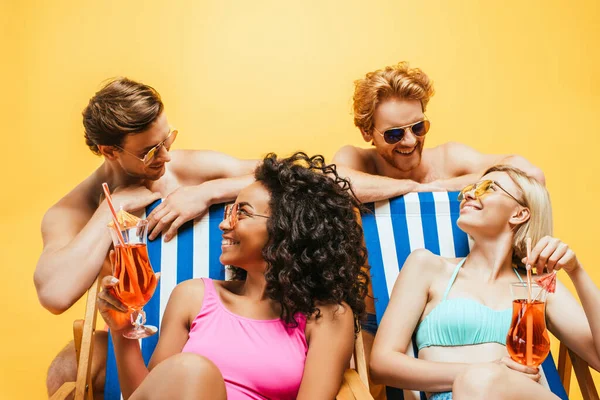 Mujeres multiculturales sentadas en tumbonas con cócteles cerca de hombres sin camisa en gafas de sol aisladas en amarillo — Stock Photo