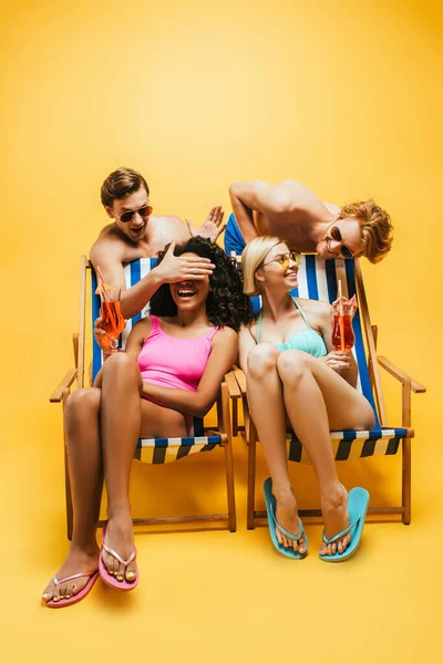 Jeunes hommes s'amusant avec des femmes multiculturelles assis sur des chaises longues avec des verres à cocktail sur jaune — Photo de stock