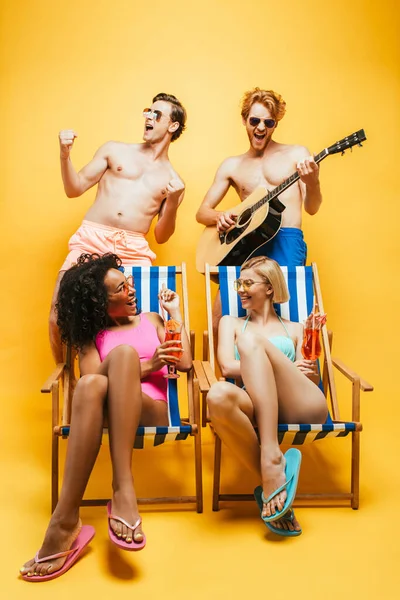 Donne multiculturali sedute su sedie a sdraio con cocktail vicino amici a torso nudo che suonano la chitarra e cantano sul giallo — Foto stock