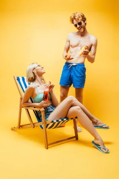 Hombre joven en pantalones cortos azules sosteniendo protector solar cerca de mujer rubia sentada en la silla de cubierta con copa de cóctel en amarillo - foto de stock