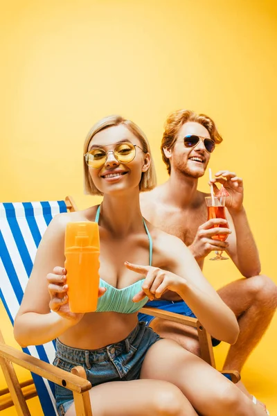 Femme blonde assise sur une chaise longue et pointant du doigt le jus d'orange près du jeune homme avec verre à cocktail isolé sur jaune — Photo de stock