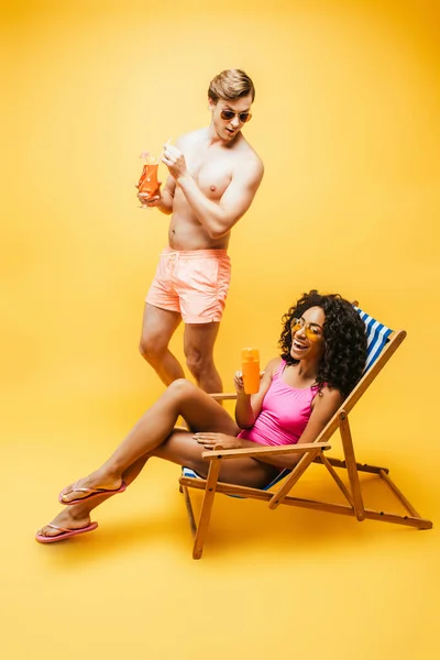 Молода американка, що сидить на палубному стільці з апельсиновим соком біля чоловіка без сорочки з коктейлем на жовтому. — стокове фото
