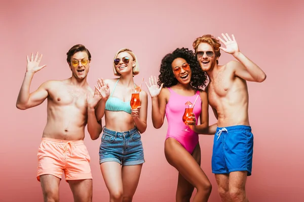 Jóvenes amigos multiculturales en traje de verano agitando las manos a la cámara mientras sostiene vasos de cóctel aislados en rosa - foto de stock