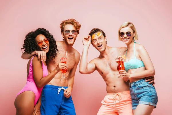 Aufgeregter Mann mit Sonnenbrille in der Nähe multikultureller Freunde, die Cocktails in der Hand halten und isoliert auf rosa Kamera schauen — Stockfoto