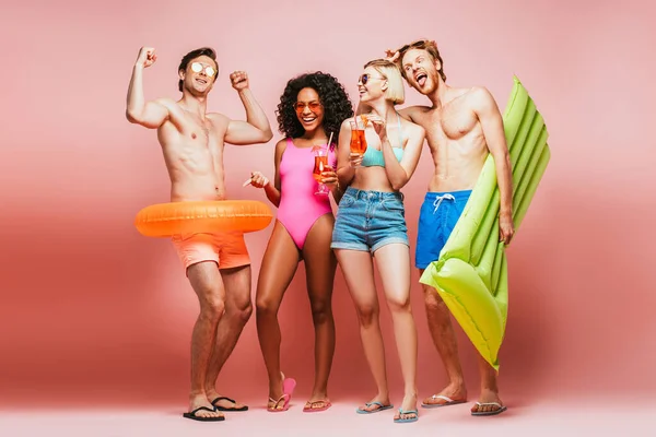 Full length view of man demonstrating biceps near multicultural friends with inflatable pool, swim ring and cocktail glasses isolated on pink — Stock Photo