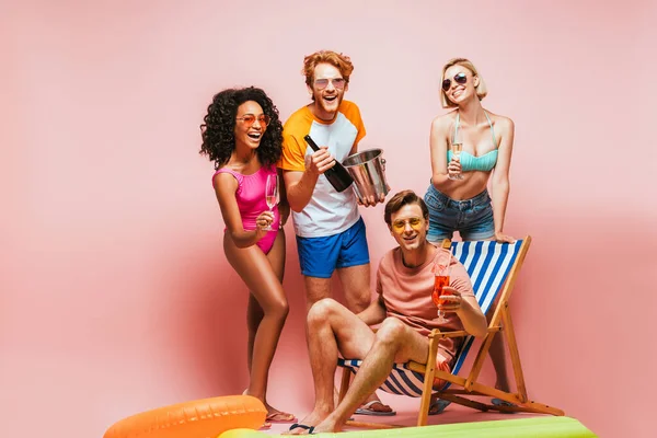 Excited multicultural friends holding champagne bottle, bucket and glasses near man sitting in deck chair with cocktail on pink — Stock Photo