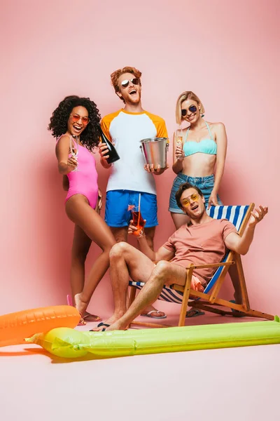 Young man sitting in deck chair with cocktail near excited multicultural friends holding champagne glasses, bottle and bucket on pink — Stock Photo