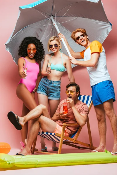 Giovane che beve cocktail sulla sedia a sdraio vicino ad amici multiculturali che tengono ombrellone e bicchieri di champagne sul rosa — Foto stock