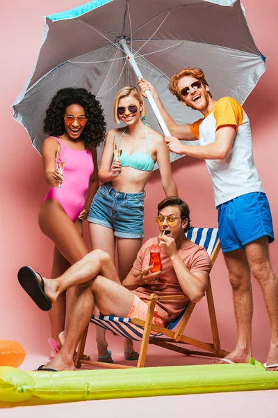 Joven bebiendo cóctel en la silla de cubierta cerca de amigos multiculturales sosteniendo sombrilla y copas de champán en rosa - foto de stock