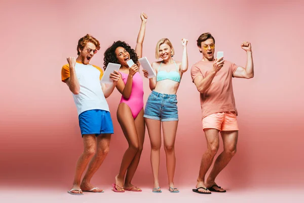 Full length view of excited multicultural friends in summer outfit showing winner gesture while looking at gadgets on pink — Stock Photo