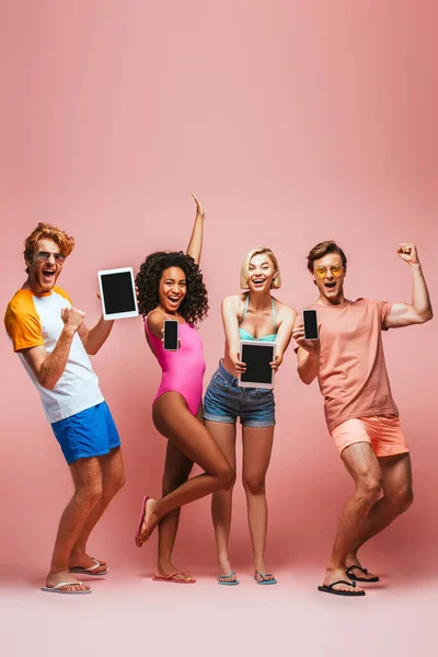 Amigos multiculturales emocionados en traje de verano mostrando gesto ganador mientras sostiene gadgets con pantalla en blanco en rosa - foto de stock