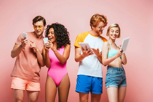 Jóvenes amigos multiculturales en traje de verano utilizando dispositivos digitales en rosa - foto de stock