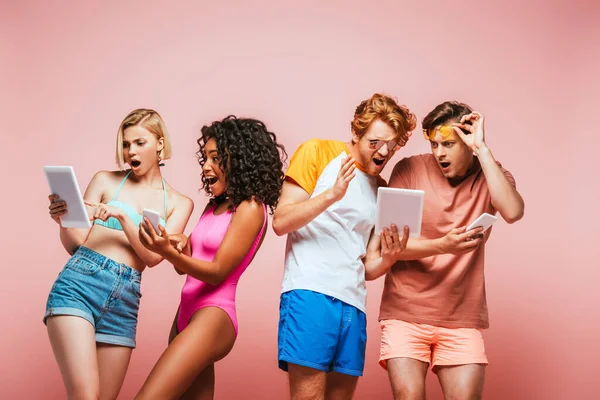 Schockierte multikulturelle Freunde im Sommeroutfit mit digitalen Geräten isoliert auf rosa — Stockfoto