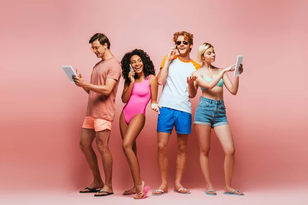 Full length view of young multicultural friends in summer outfit using digital devices on pink — Stock Photo