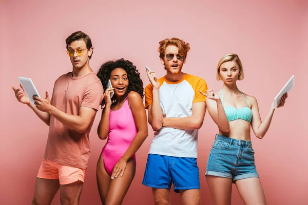 Мультиэтнические друзья в купальниках и солнцезащитных очках держат смартфоны и цифровые планшеты на розовом фоне — стоковое фото