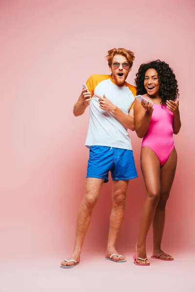 Pareja multicultural en traje de baño y gafas de sol apuntando a teléfonos inteligentes sobre fondo rosa - foto de stock