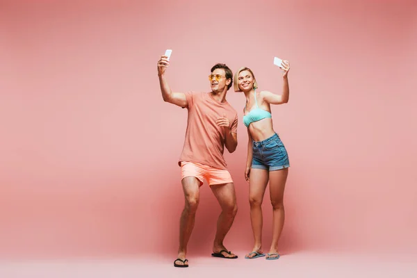 Jeune couple en lunettes de soleil et maillot de bain prenant selfie avec smartphones sur fond rose — Photo de stock