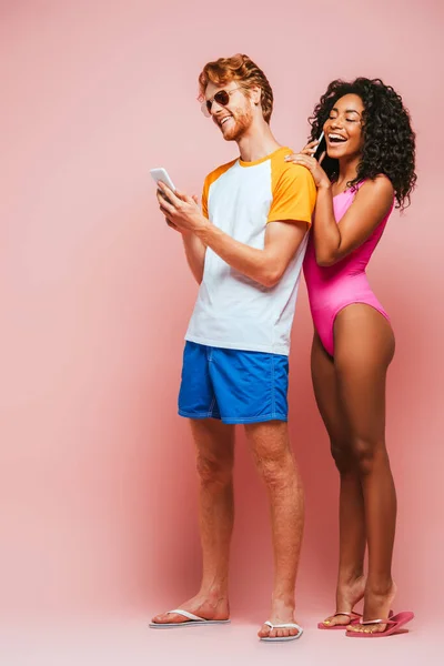 Afroamerikanerin im Badeanzug spricht auf Smartphone neben Mann mit Sonnenbrille auf rosa Hintergrund — Stockfoto