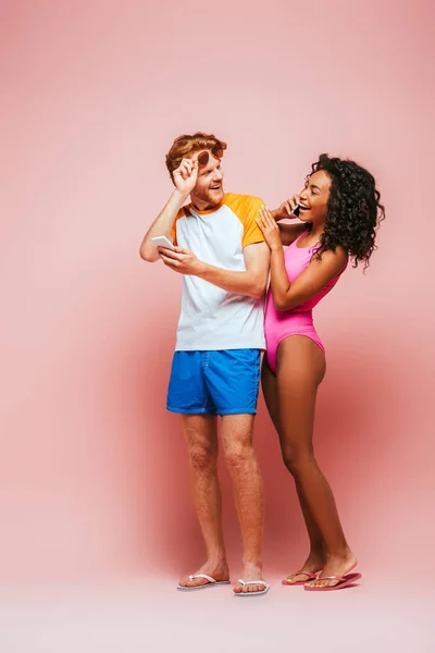 Hombre sosteniendo teléfono inteligente y gafas de sol mientras mira a la mujer afroamericana en traje de baño hablando en el teléfono inteligente sobre fondo rosa - foto de stock