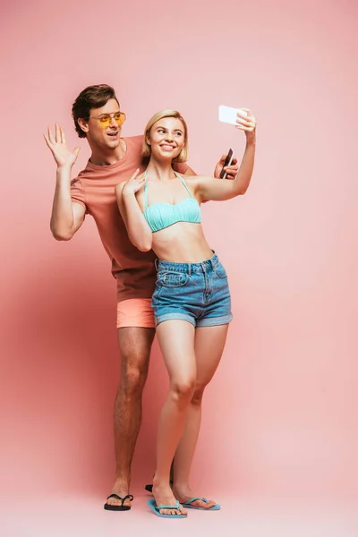 Homme en lunettes de soleil ayant appel vidéo près de la femme en maillot de bain tenant smartphone sur fond rose — Photo de stock