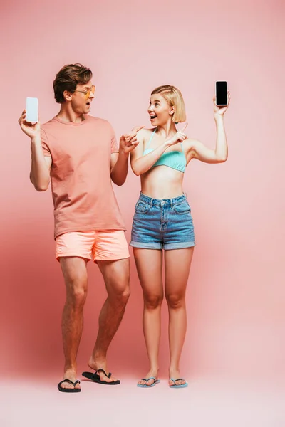 Young friends in flip flops and swimsuit pointing with fingers at smartphone with blank screen on pink background — Stock Photo