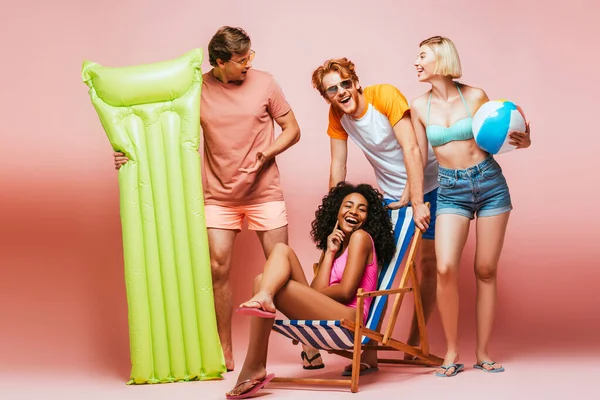 Yong multiethnische Freunde in Badeanzügen und Sonnenbrille mit aufblasbarem Ball und Matratze auf rosa Hintergrund — Stockfoto