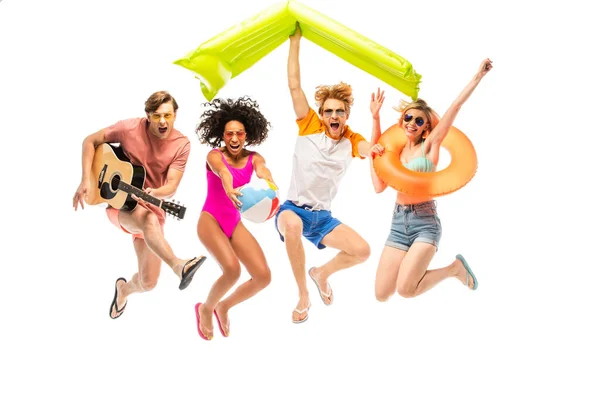 Emocionados amigos multiétnicos con bola inflable, colchón y guitarra acústica saltando aislados en blanco - foto de stock
