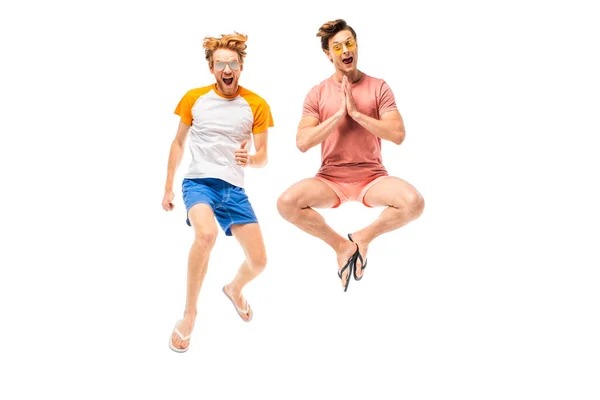 Amigos emocionados en gafas de sol que muestran por favor gesto y pulgar hacia arriba mientras salta aislado en blanco - foto de stock