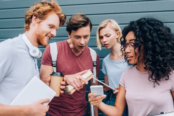 Étudiants multiethniques concentrés utilisant un smartphone tout en tenant des livres dans la rue urbaine — Photo de stock