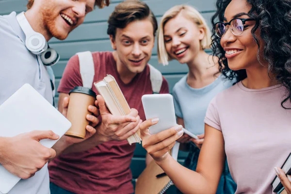 Focus selettivo di studenti multiculturali che utilizzano smartphone mentre tengono libri e caffè per andare per strada urbana — Foto stock