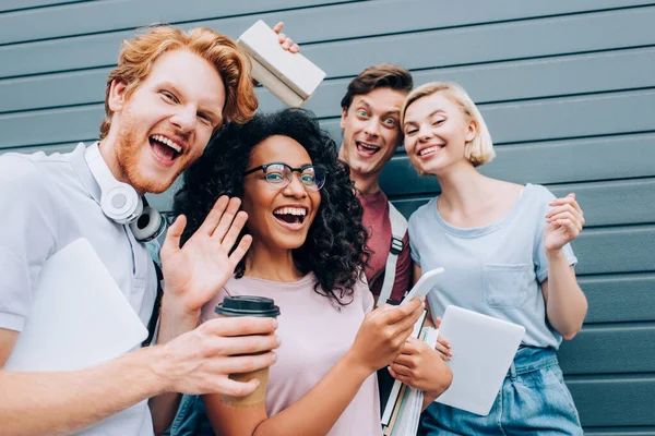 Enfoque selectivo de estudiantes multiculturales con café para llevar y dispositivos digitales saludando con la mano a la cámara en la calle urbana - foto de stock