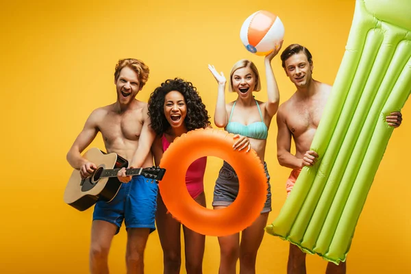 Multiethnic friends with acoustic guitar, inflatable mattress and ball on yellow background — Stock Photo