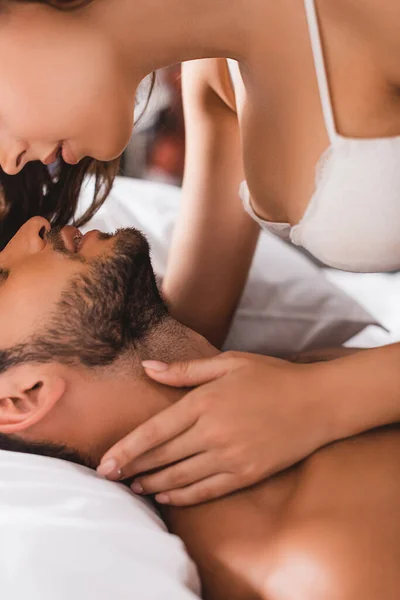Vista recortada de la mujer sexy en sujetador tocando el cuello del hombre muscular en la cama - foto de stock
