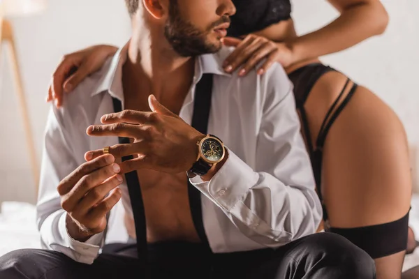 Cropped view of man in shirt taking off wedding ring near sensual woman on bed — Stock Photo