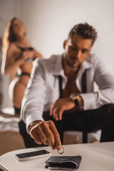Селективный фокус мужчины положить обручальное кольцо возле бумажника и смартфона на кофейный столик рядом с женщиной в нижнем белье на кровати — стоковое фото