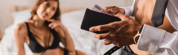Panoramic shot of muscular man holding wallet and money near woman in bra on bed — Stock Photo