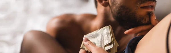 Panoramic crop of woman holding dollars and touching chin of shirtless man on bed — Stock Photo