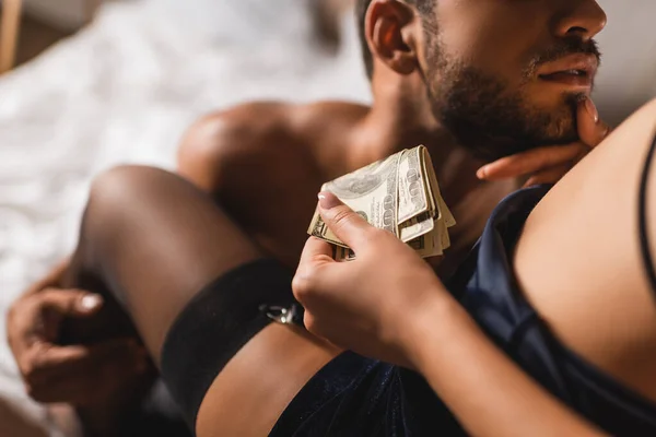 Vista cortada de mulher sexy segurando notas de dólar e tocando queixo de homem sem camisa no quarto — Fotografia de Stock