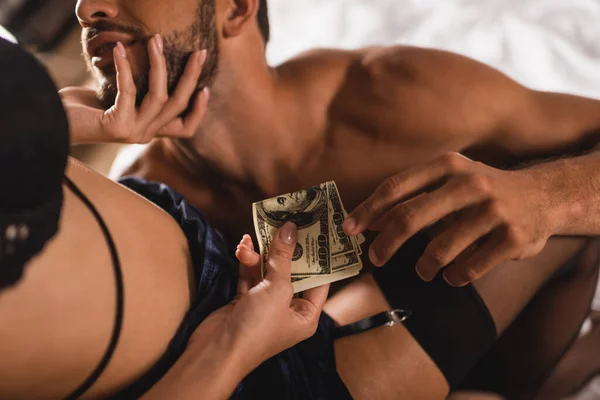 Vista recortada de mujer seductora en sujetador y media dando dólares y tocando al hombre musculoso en la cama - foto de stock