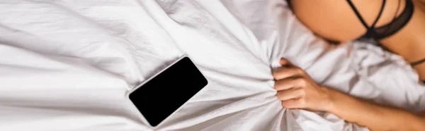 Imagen panorámica de smartphone con pantalla en blanco sobre ropa de cama cerca de mujer sexy en lencería en la cama - foto de stock