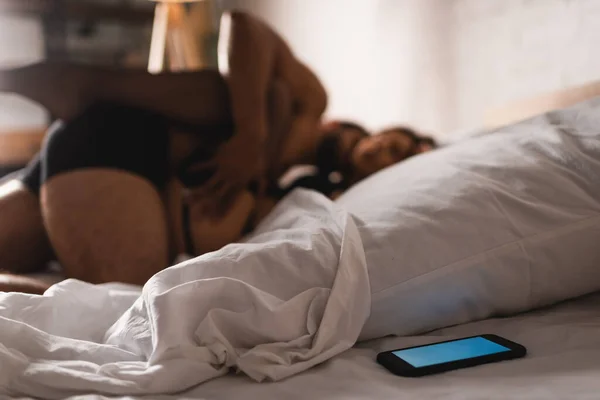 Selective focus of smartphone near pillow and sexy couple lying on bed at background — Stock Photo