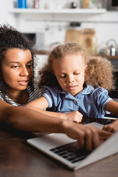Селективный фокус африканской американки, указывающей на ноутбук рядом с концентрированной дочерью — стоковое фото