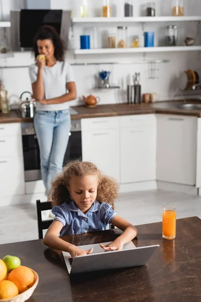 Concentrée afro-américaine fille à l'aide d'un ordinateur portable tout en étant assis à la table de cuisine près de mère debout sur le fond — Photo de stock