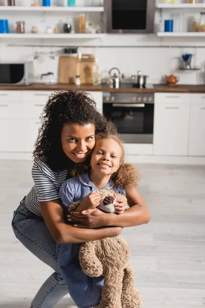 Young african american woman embracing excited daughter with teddy bear while looking at camera in kitchen — Stock Photo