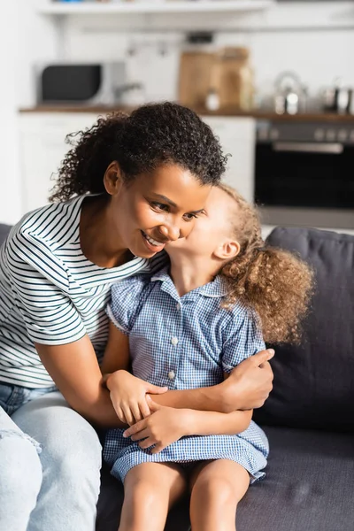 African american girl whispering in ear of mother while sitting on sofa at home — Stock Photo
